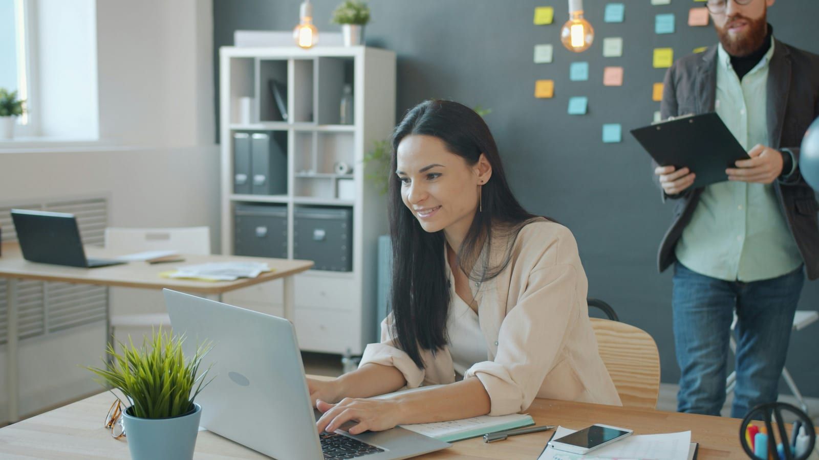 A smiling woman working on a laptop at a bright office desk beside a potted plant
