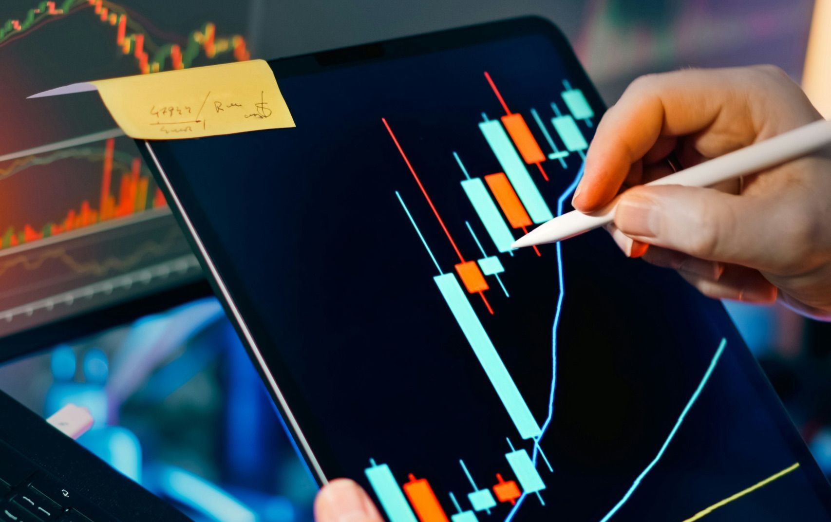 A person marking up a candlestick chart on a tablet at a desk of trading screens