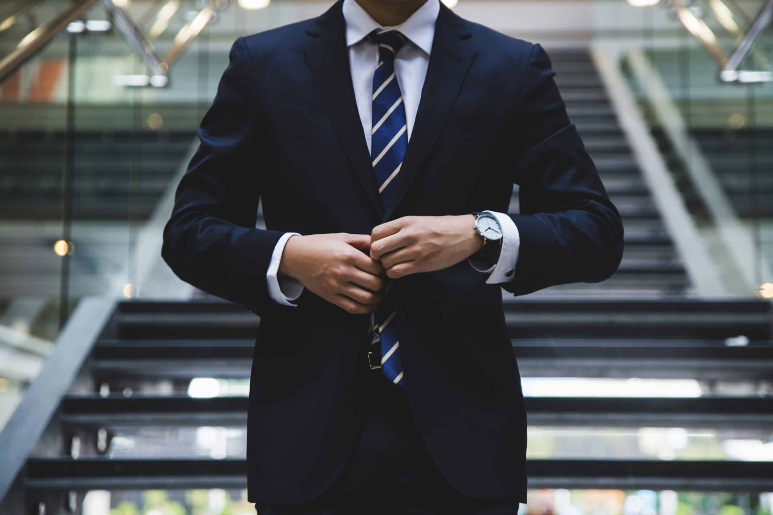 The body of a man wearing a dark blue suit with a blurred office background.