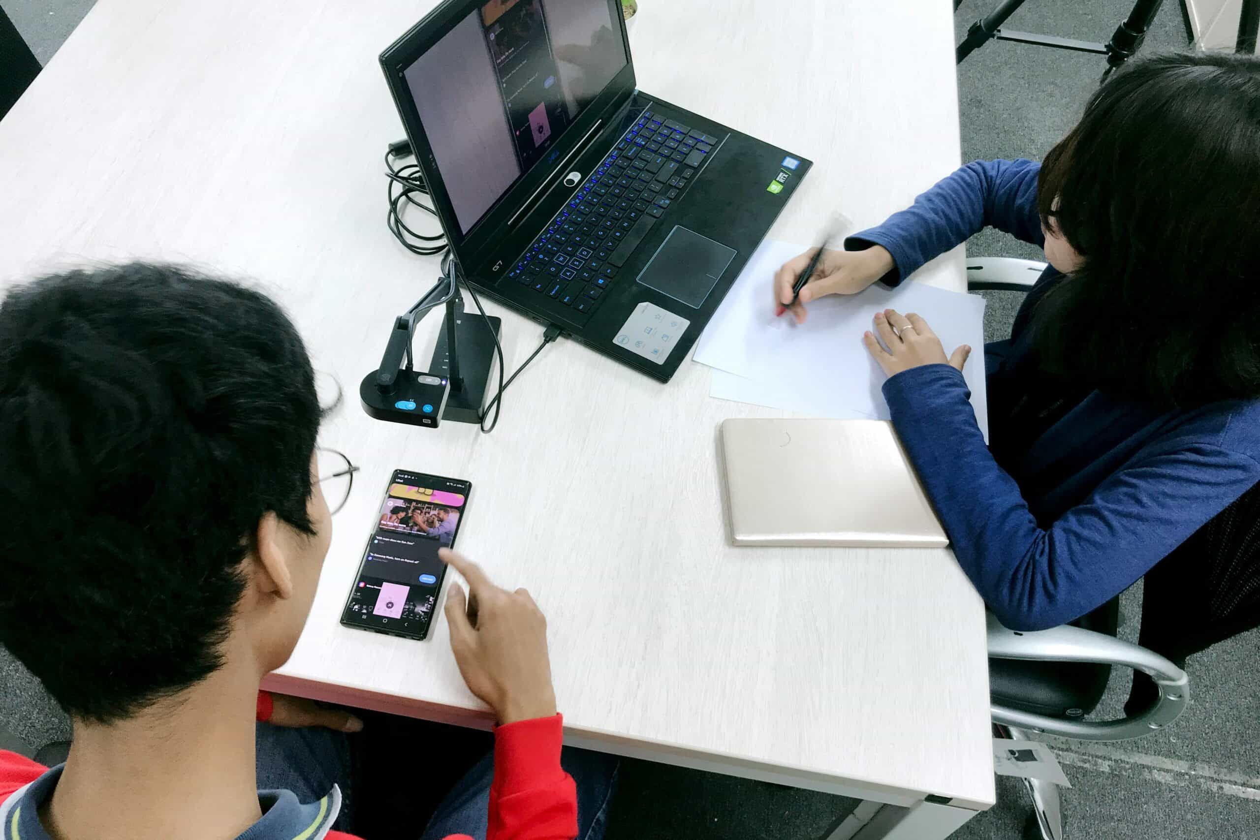 A man with a mobile phone and a woman with a laptop are working together in an office around a table.